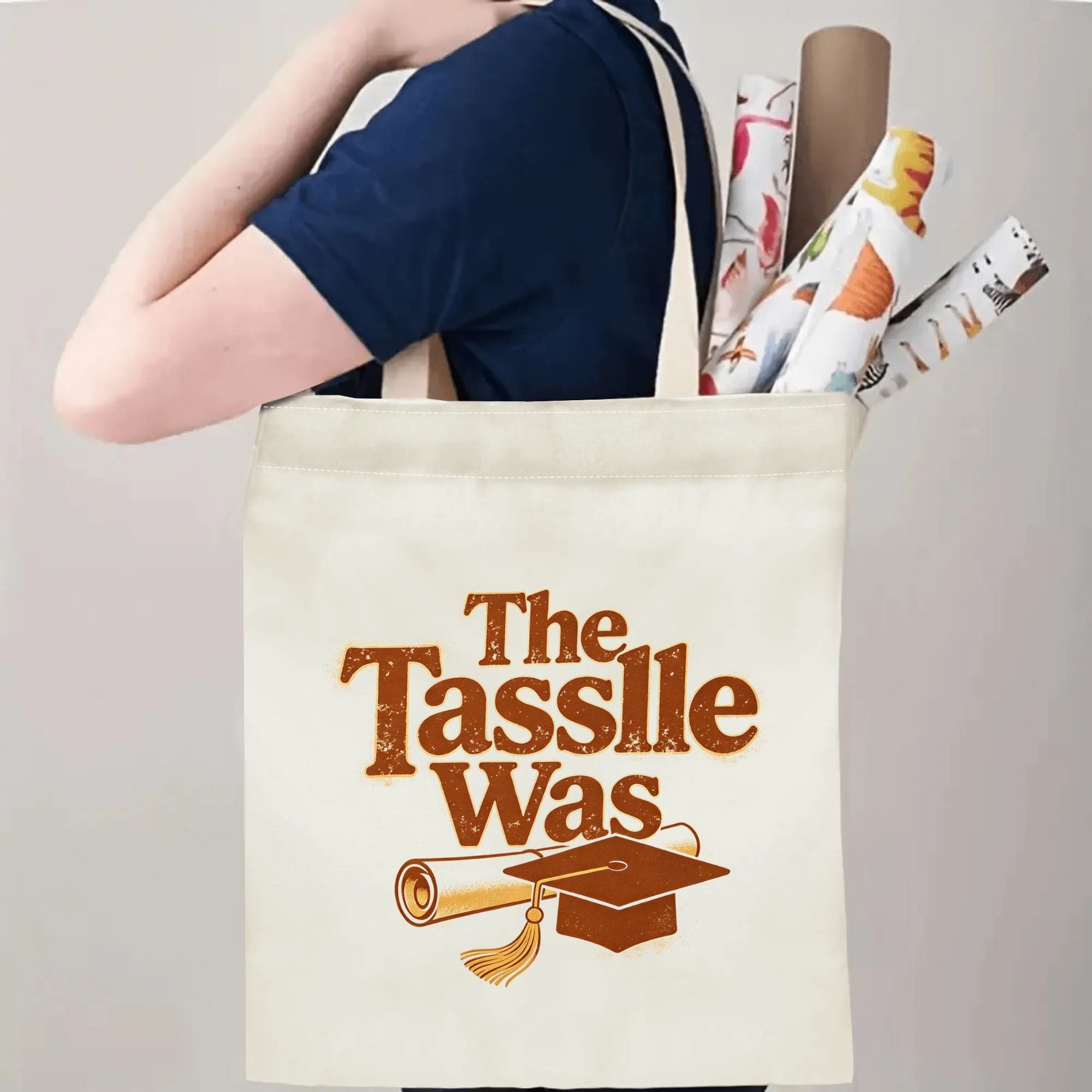 Person carrying a “The Tassle Was” graduation tote bag over the shoulder, with rolled posters and graduation essentials.