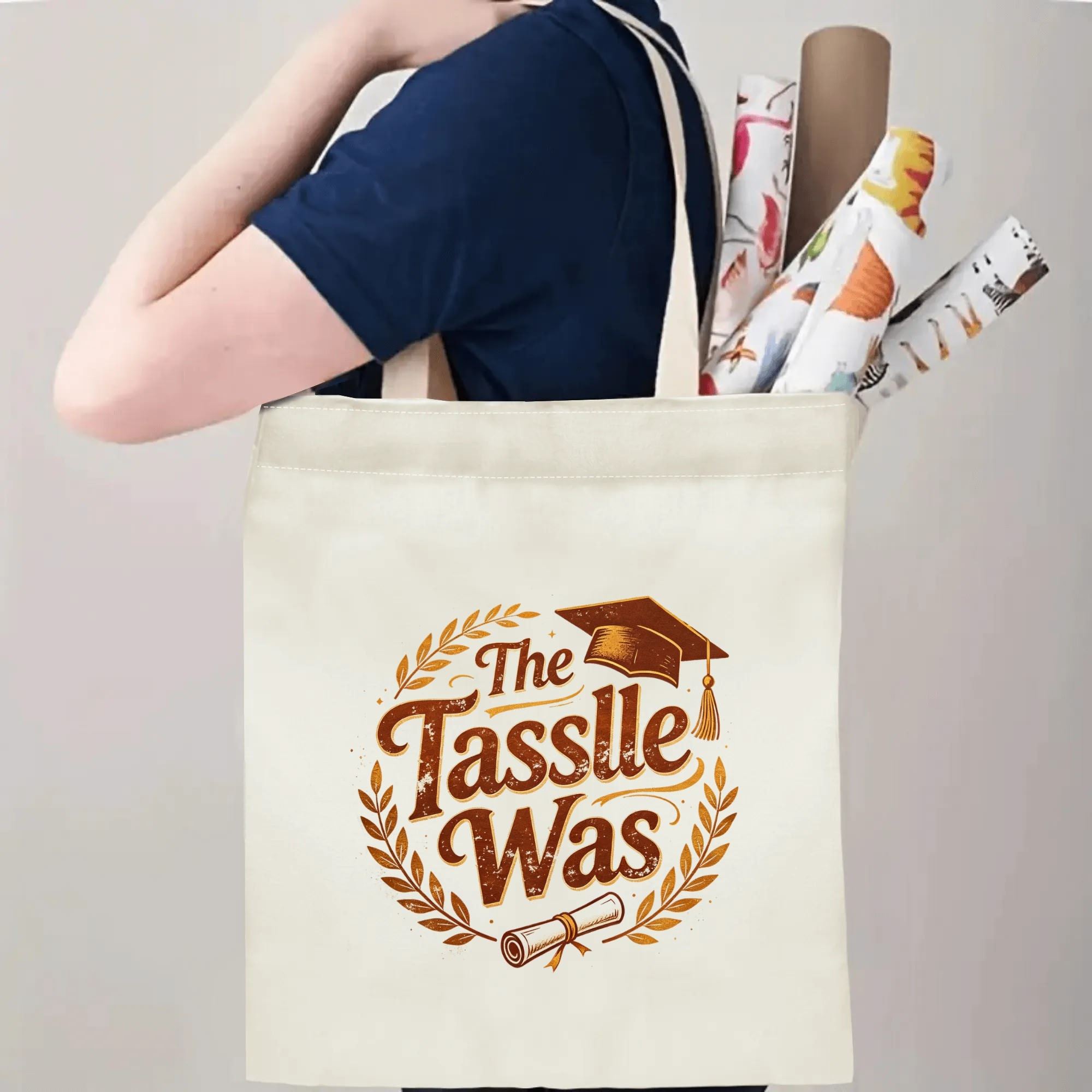 Person carrying a “The Tassle Was” graduation tote bag over the shoulder, with rolled posters and graduation essentials.