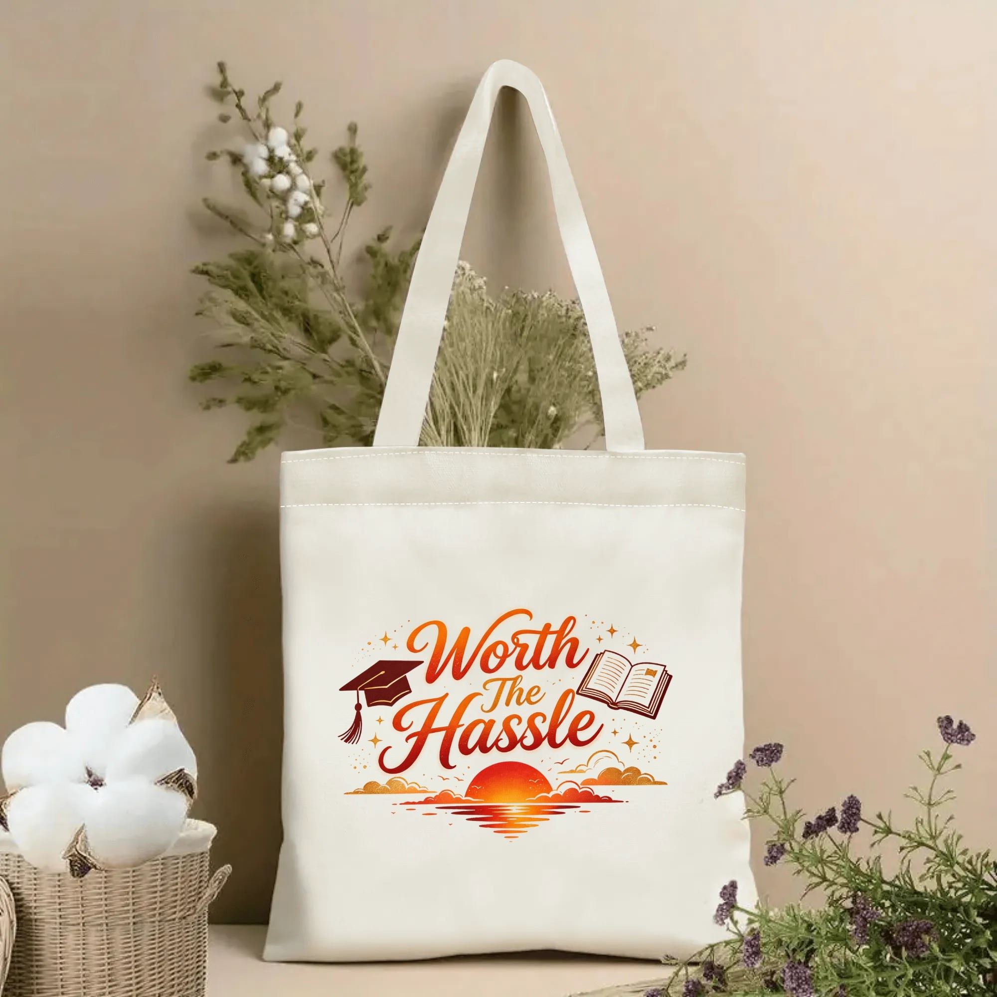 Natural canvas tote bag with “Worth The Hassle” print, featuring a graduation cap, open book, sunset scene, and star accents.