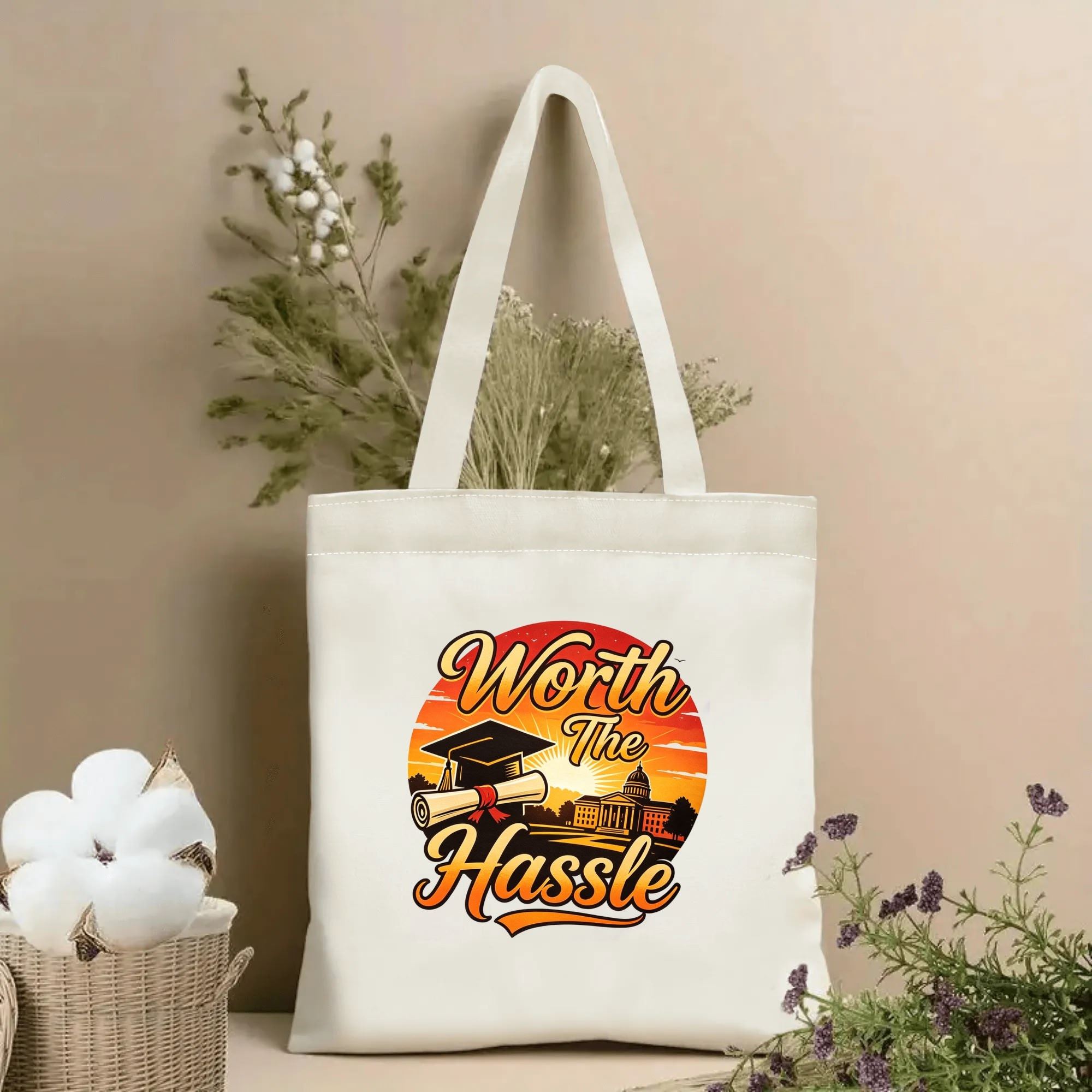 Natural canvas tote bag with “Worth The Hassle” print, featuring a graduation cap, diploma, university building, and sunset scene.
