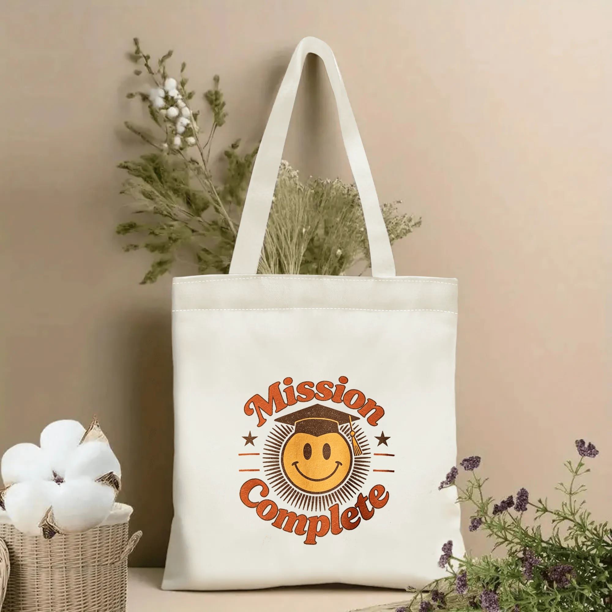 Natural canvas tote bag with “Mission Complete” print, featuring a smiley face wearing a graduation cap, surrounded by a retro sunburst and star accents.