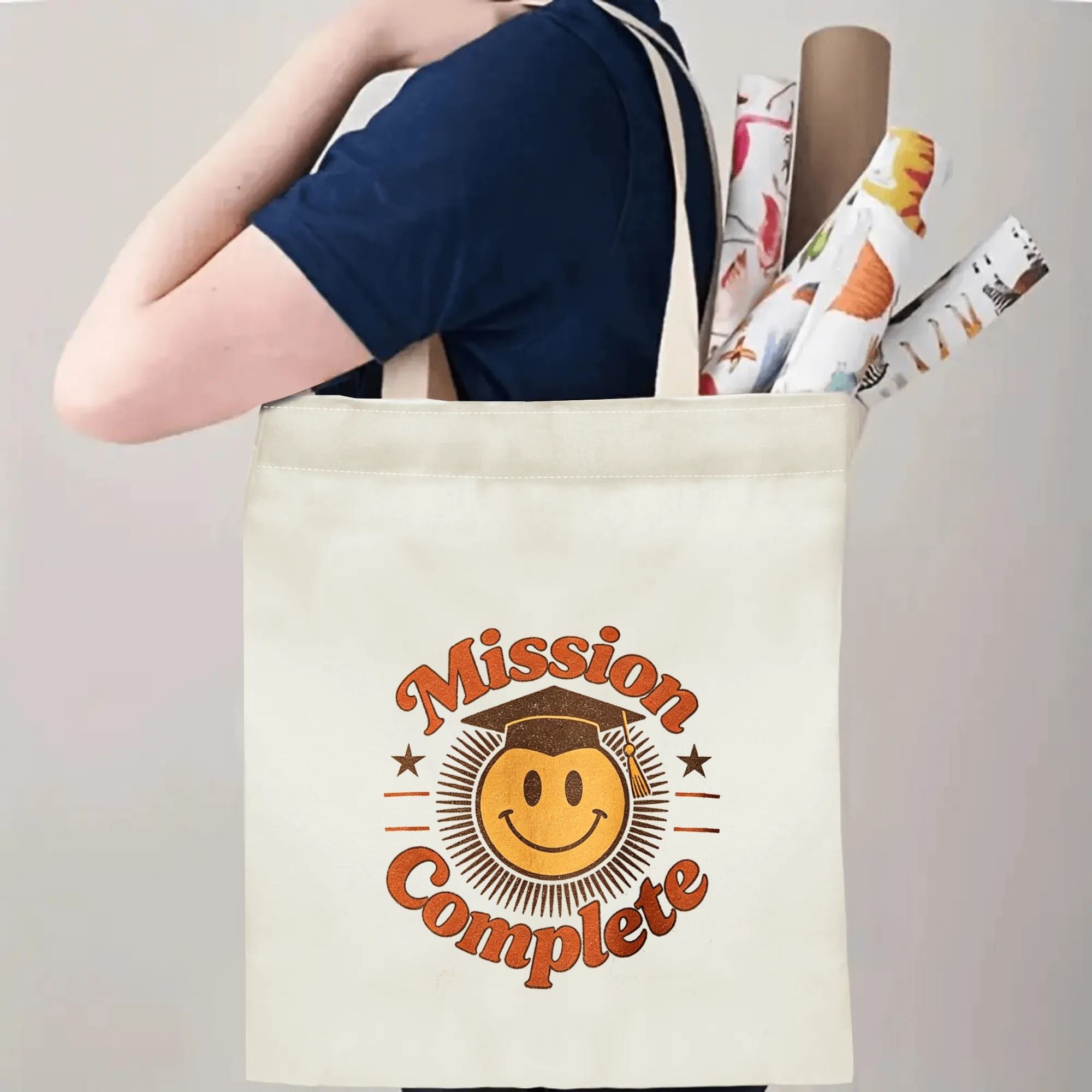 Person carrying a “Mission Complete” graduation tote bag over the shoulder, with rolled posters and graduation essentials.