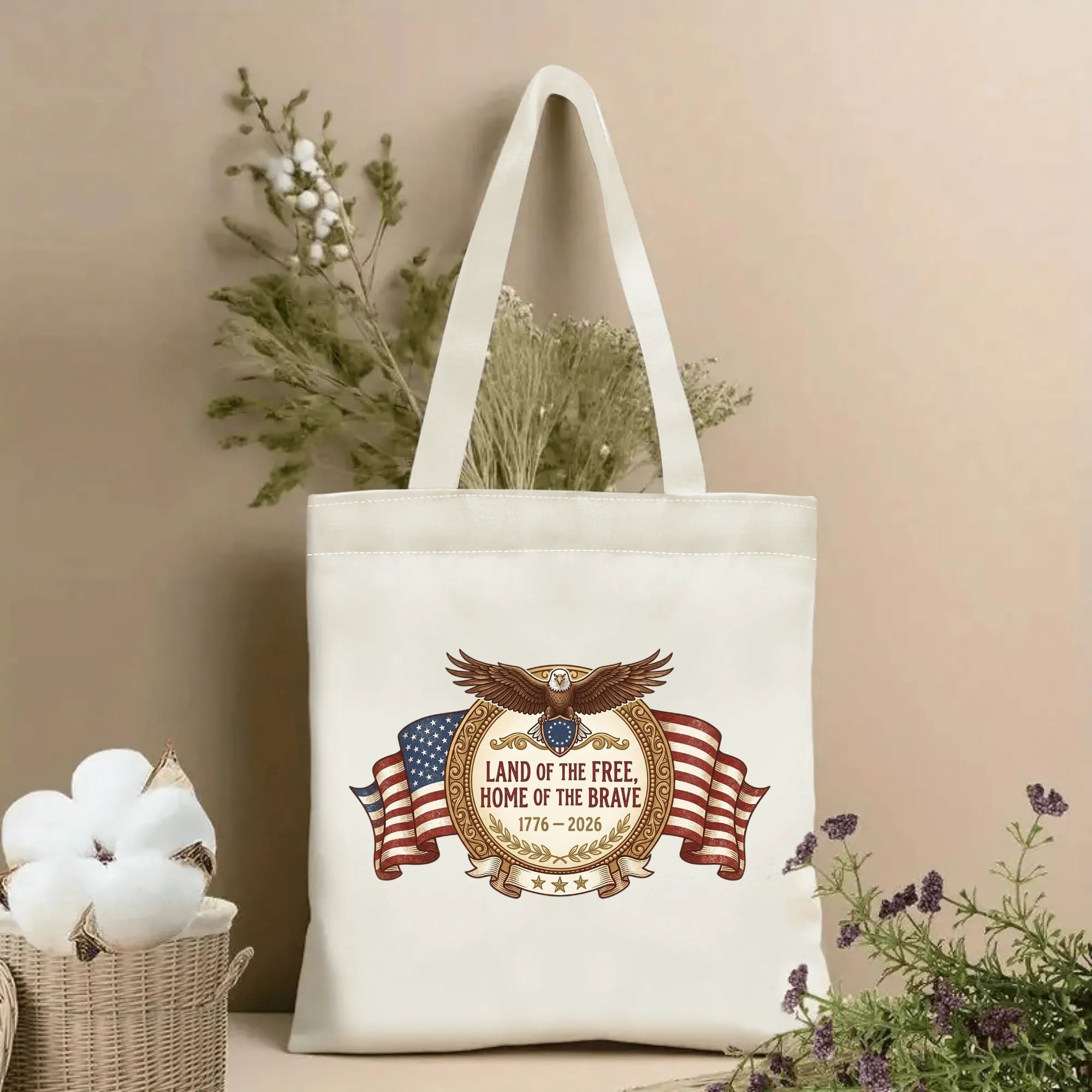 Beige reusable canvas tote decorated with vintage bald eagle & American flag scroll print "Land of the Free Home of the Brave 1776-2026", displayed beside dried botanicals.