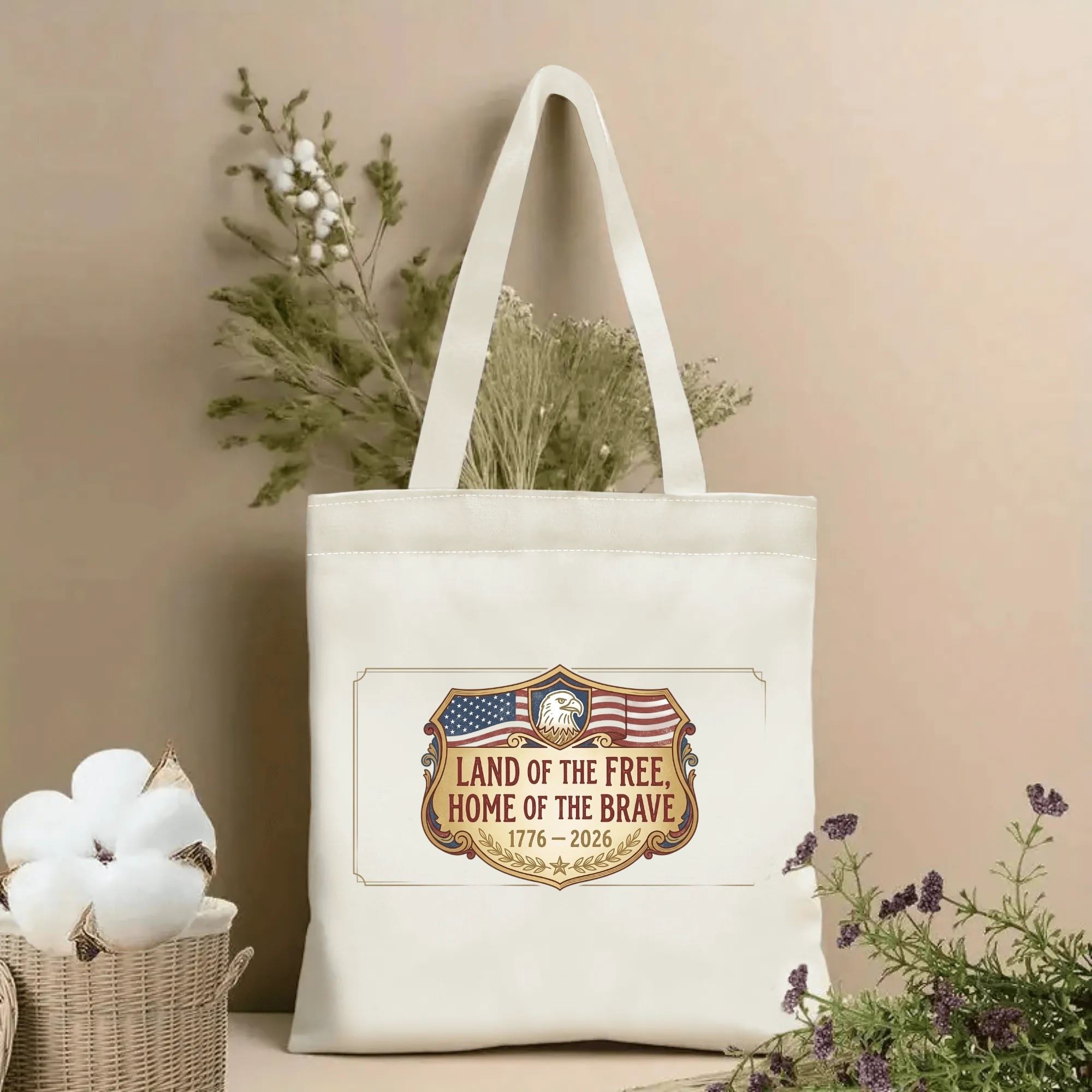 Beige reusable canvas tote bag with vintage shield-style bald eagle & USA flag print and “Land of the Free Home of the Brave 1776-2026” patriotic slogan, displayed beside dried floral decor.