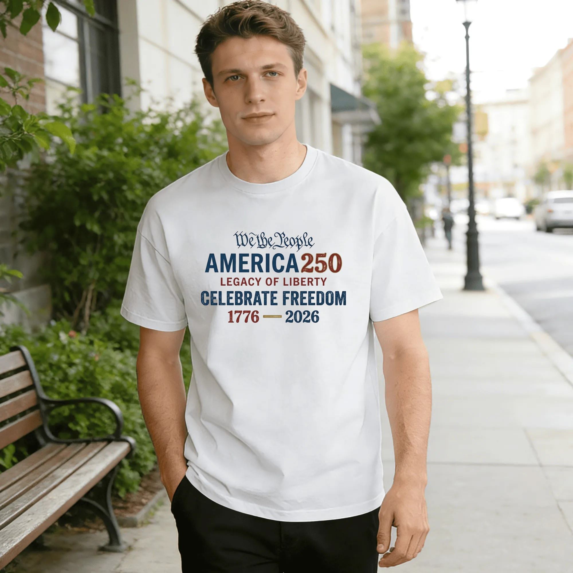 Young man wearing white relaxed‑fit cotton tee with clean stacked vintage‑text America 250 graphic, “We The People” script, “America 250” bold lettering, “Legacy of Liberty” & “Celebrate Freedom” mottos, 1776‑2026 anniversary dates, distressed red‑white‑blue typography, street‑style outdoor lifestyle shot, simple classic USA 250th‑anniversary commemorative apparel
