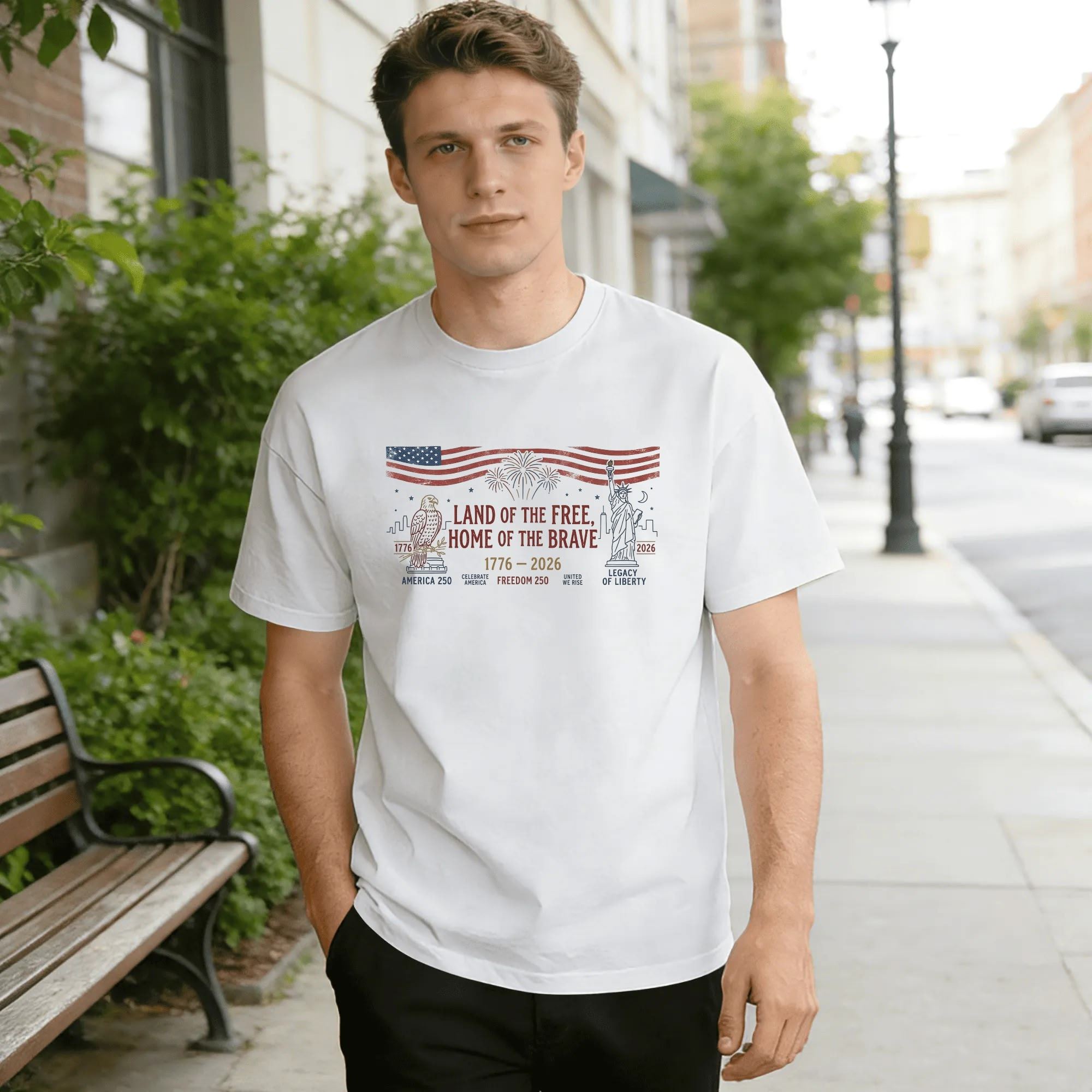 Young man wearing white relaxed‑fit cotton tee with retro distressed America 250 graphic, top waving US flag, central “Land of the Free, Home of the Brave” text, side bald eagle & Statue of Liberty silhouettes, fireworks accents, 1776‑2026 anniversary marks, vintage worn‑finish style, street‑style outdoor lifestyle shot, casual USA 250th‑anniversary commemorative apparel