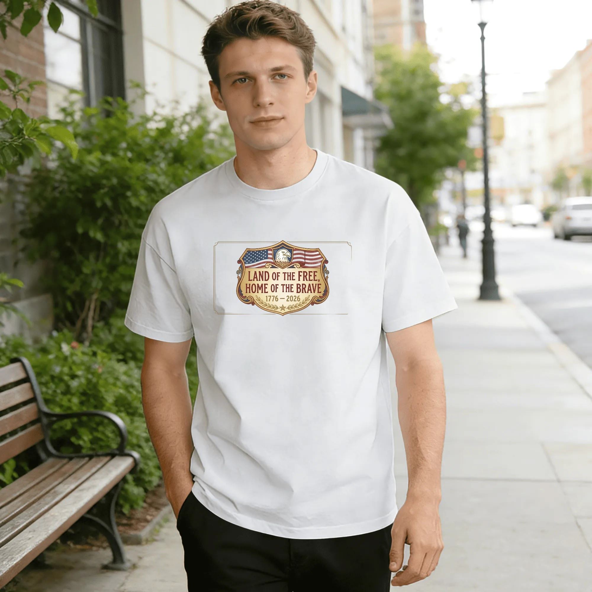 Young man wearing white relaxed‑fit cotton tee with elegant shield‑badge America 250 graphic, golden‑trimmed framed shield, top eagle head, American flag overlay, golden laurel base, “Land of the Free, Home of the Brave” slogan, 1776‑2026 anniversary dates, classic vintage emblem design, street‑style outdoor lifestyle shot, refined USA 250th‑anniversary commemorative apparel