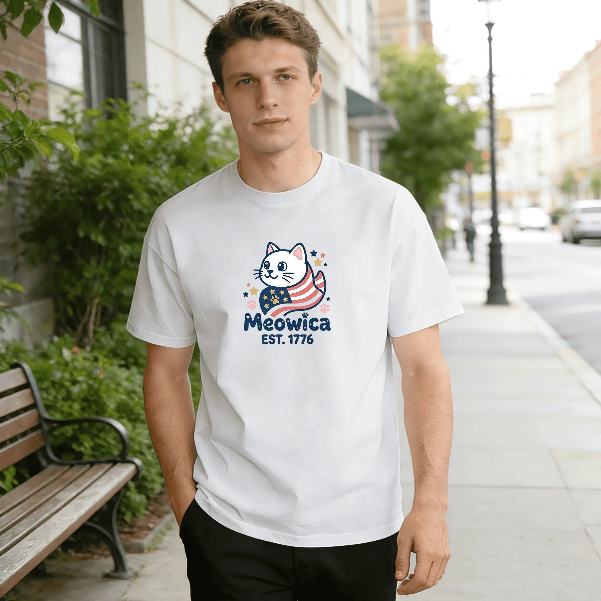 Young man wearing white relaxed‑fit cotton tee with sweet cartoon America 250 graphic, adorable cat wrapped in US flag‑striped scarf, star‑paw accents, “Meowica EST.1776” playful lettering, clean casual style, street‑style outdoor lifestyle shot, simple novelty patriotic commemorative apparel
