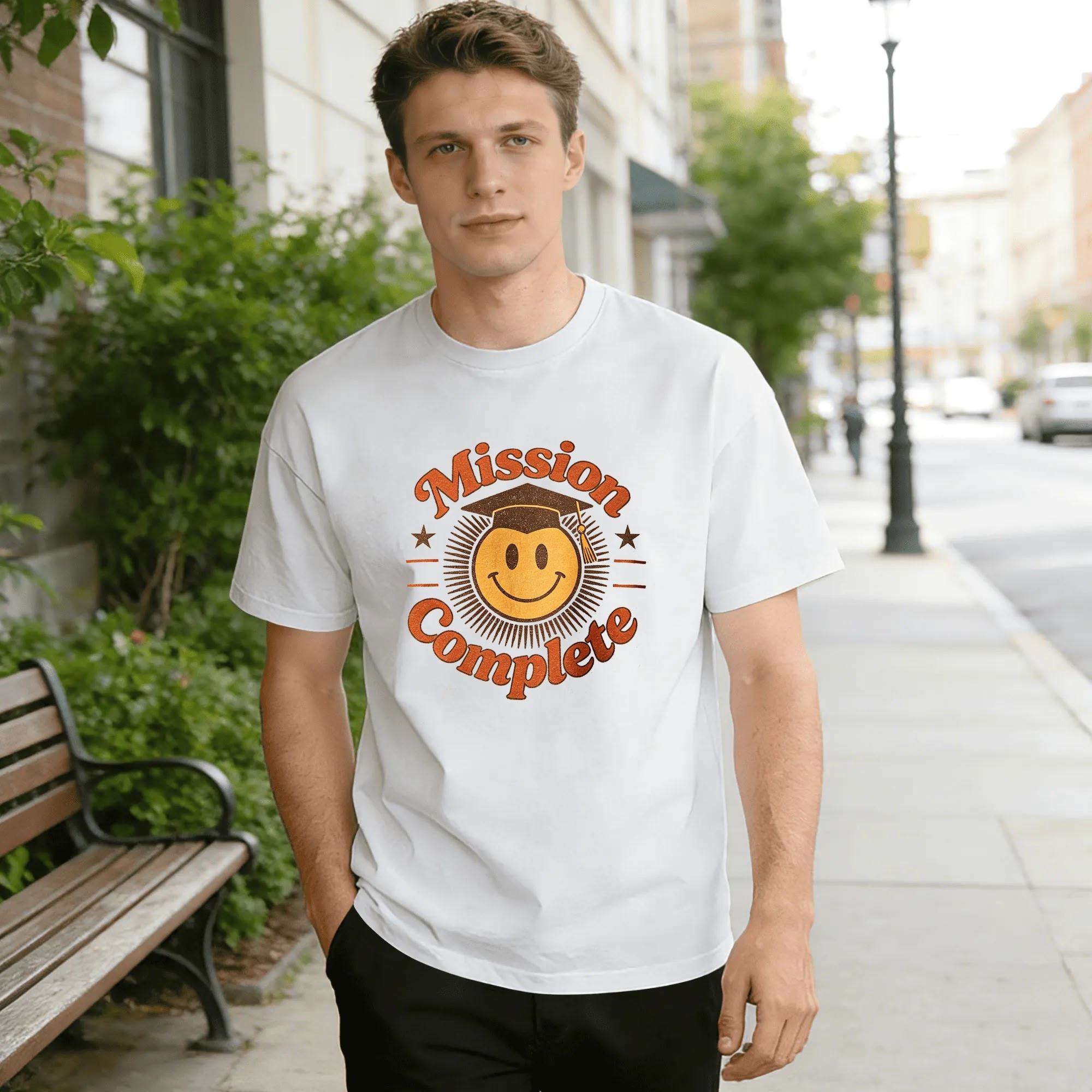 White crewneck T-shirt with a retro “Mission Complete” graphic, featuring a smiley face in a graduation cap, distressed orange lettering, and star accents.
