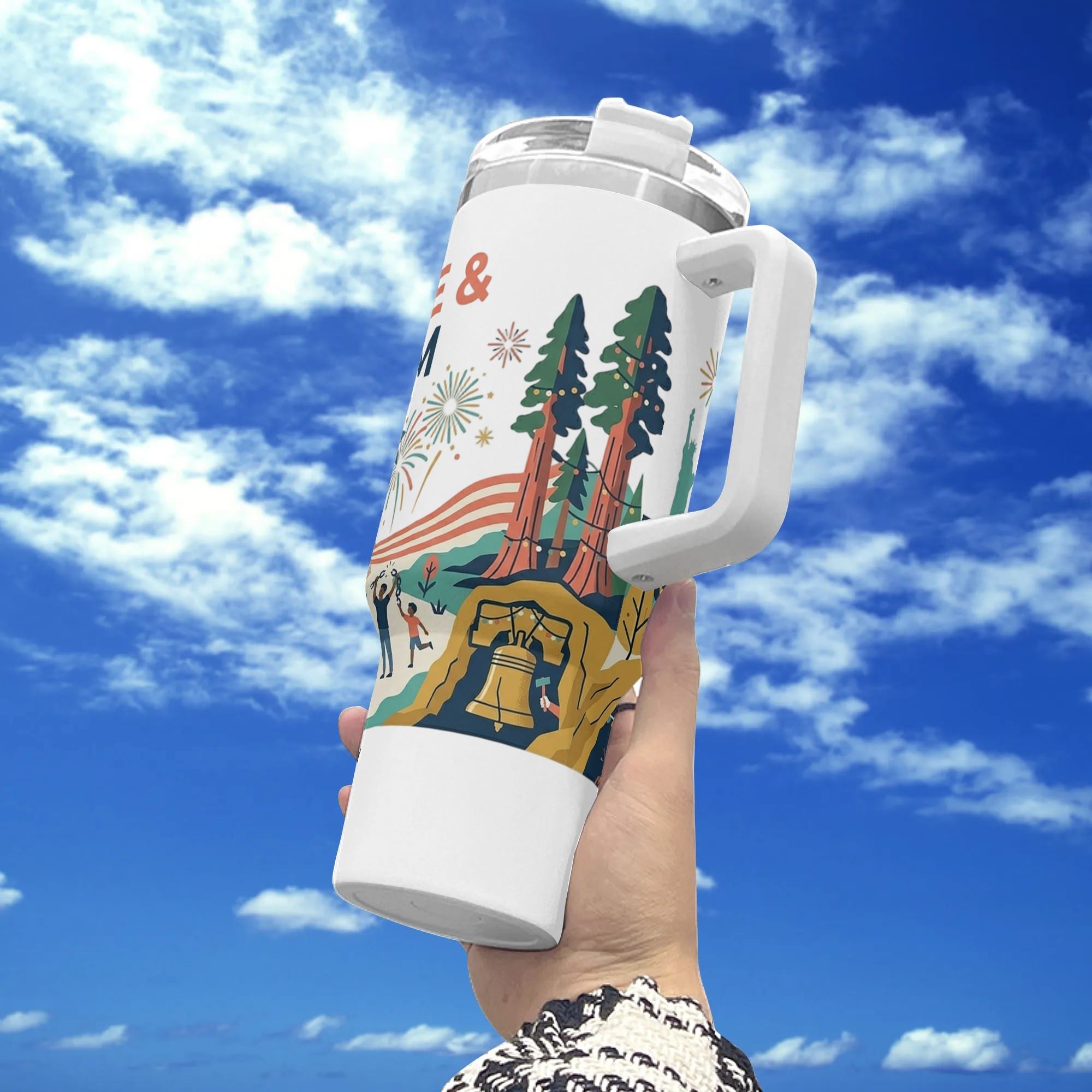 Hand-held shot outdoors against cloudy sky, white 40oz stainless steel tumbler wrapped with full-wrap vintage patriotic “Celebrate Freedom 1776-2026” print including Liberty Bell, pine trees, fireworks and festive crowd graphic.