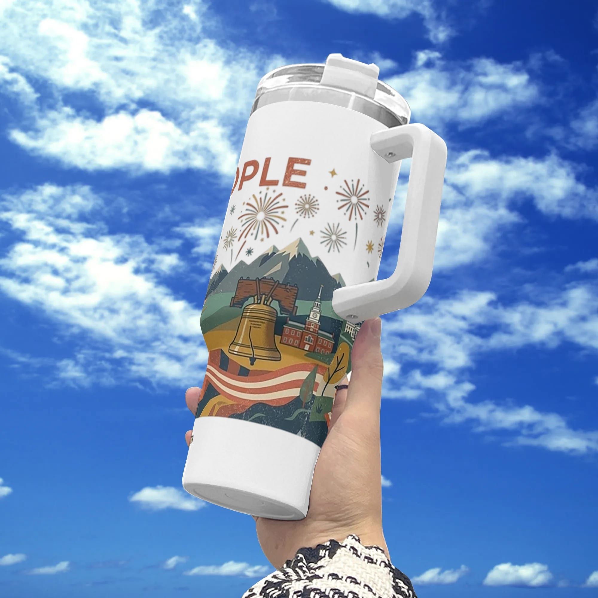 Handheld outdoor photo against blue cloudy sky, white 40oz insulated tumbler with full-wrap “We The People” patriotic print featuring Liberty Bell, Independence Hall, mountain range and festive fireworks.