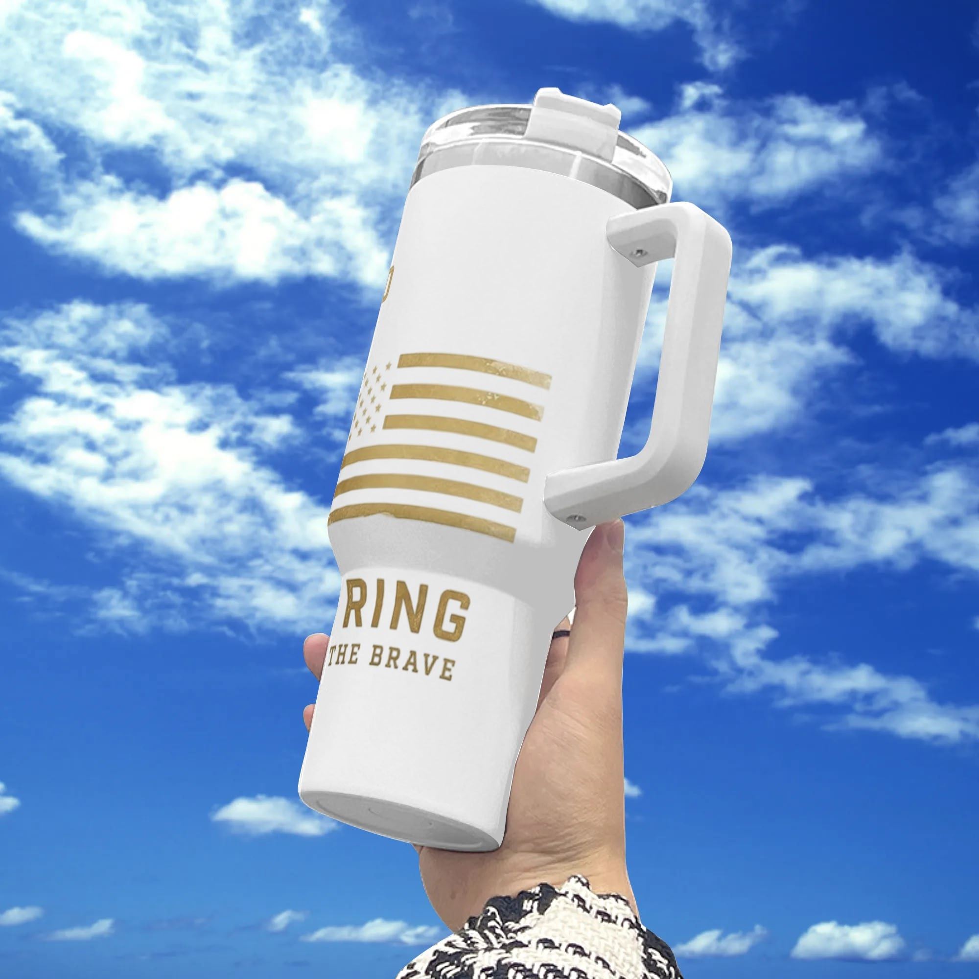 Hand-held outdoor shot under blue cloudy sky, white 40oz insulated tumbler with golden vintage print: faded gold USA flag stripes plus “HOME OF THE BRAVE” patriotic lettering.