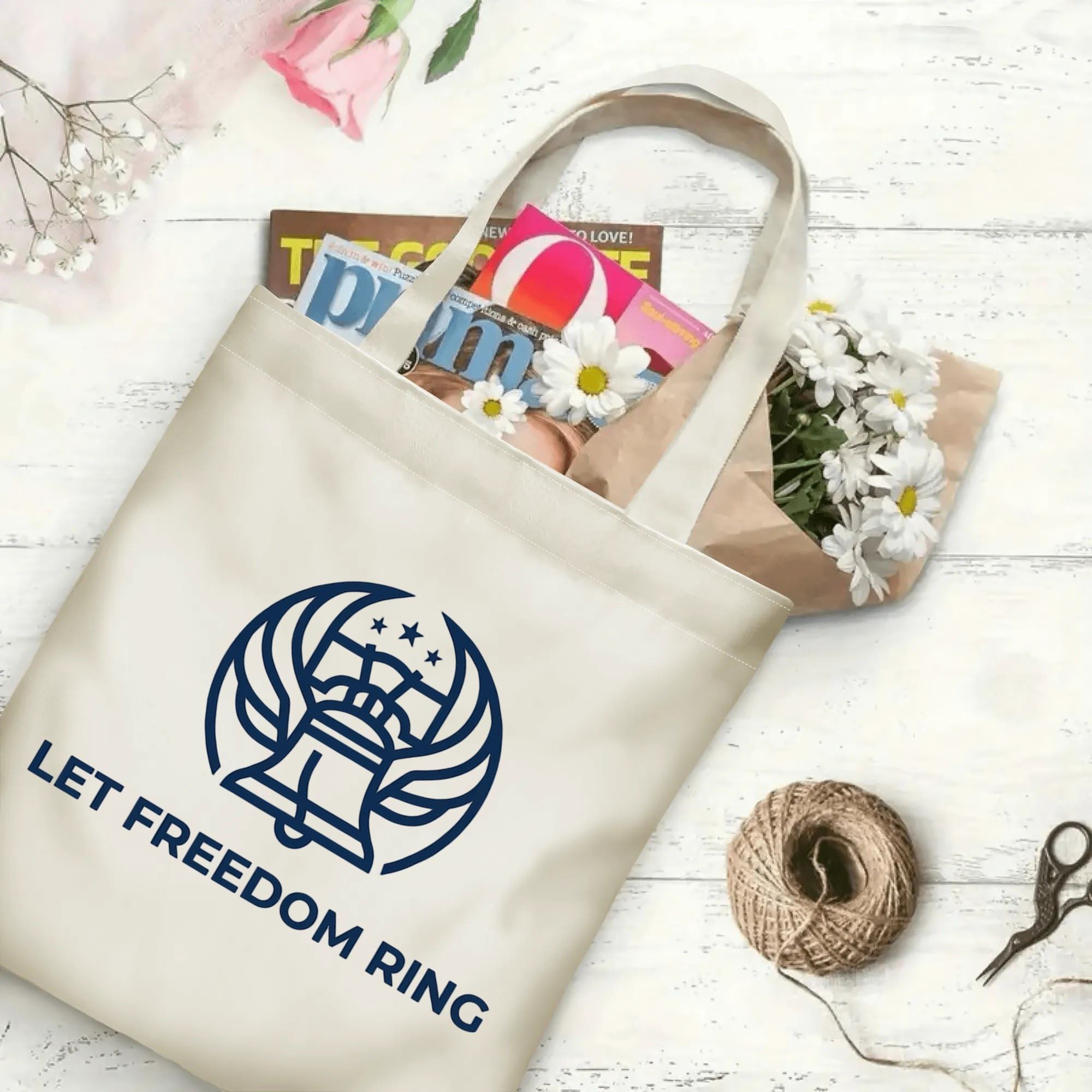 Patriotic Liberty Bell logo printed beige canvas bag laid on rustic wood with magazines and fresh daisy flowers