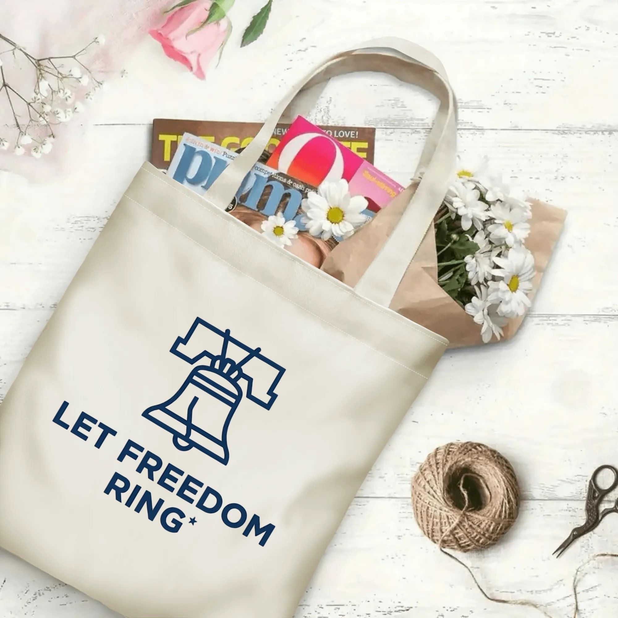 Patriotic Liberty Bell slogan printed beige canvas bag laid on rustic wooden board filled with magazines and fresh daisy blooms
