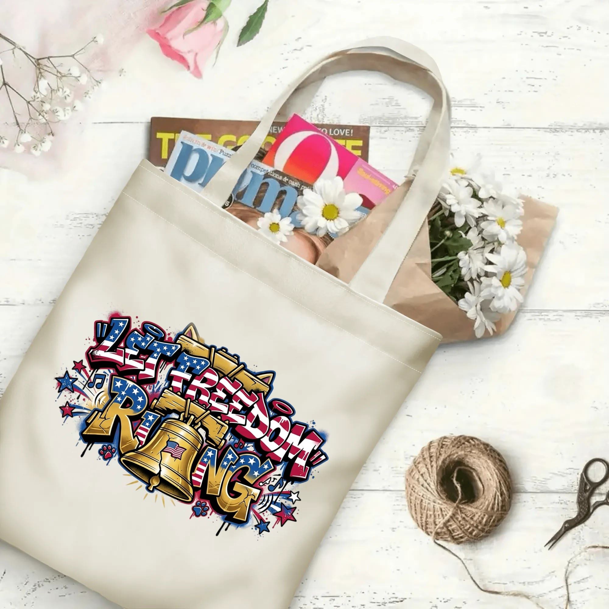 Wooden table flat lay: Patriotic street art Liberty Bell canvas tote filled with magazines and fresh daisies on rustic wood surface