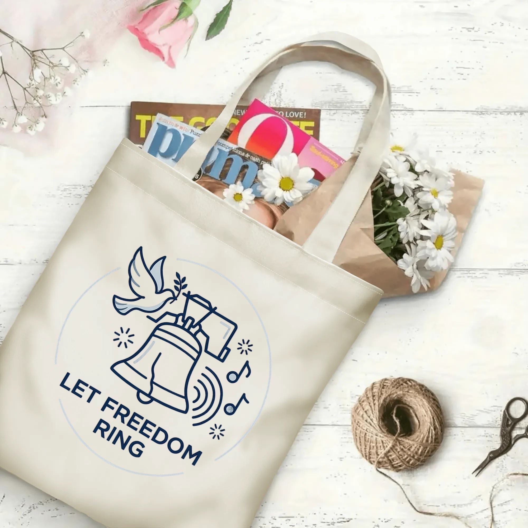 Minimalist line-drawn freedom bell & dove printed canvas tote laid on rustic wooden board with fresh daisies and magazines