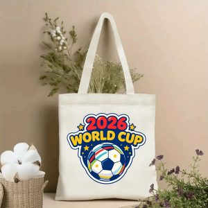 Lifestyle shot of the tote bag with a vibrant graffiti-style “2026 World Cup” print, featuring a soccer ball and colorful paint splatters, displayed against a neutral background with plants.