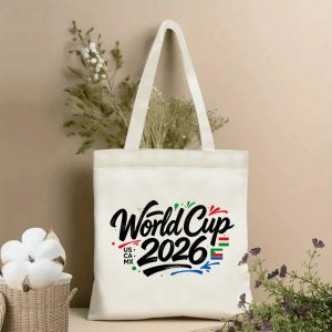 Lifestyle shot of the tote bag featuring a brush-script “World Cup 2026” print, accented with colorful splashes and the host country initials “US • CA • MX,” displayed against a neutral background with plants.