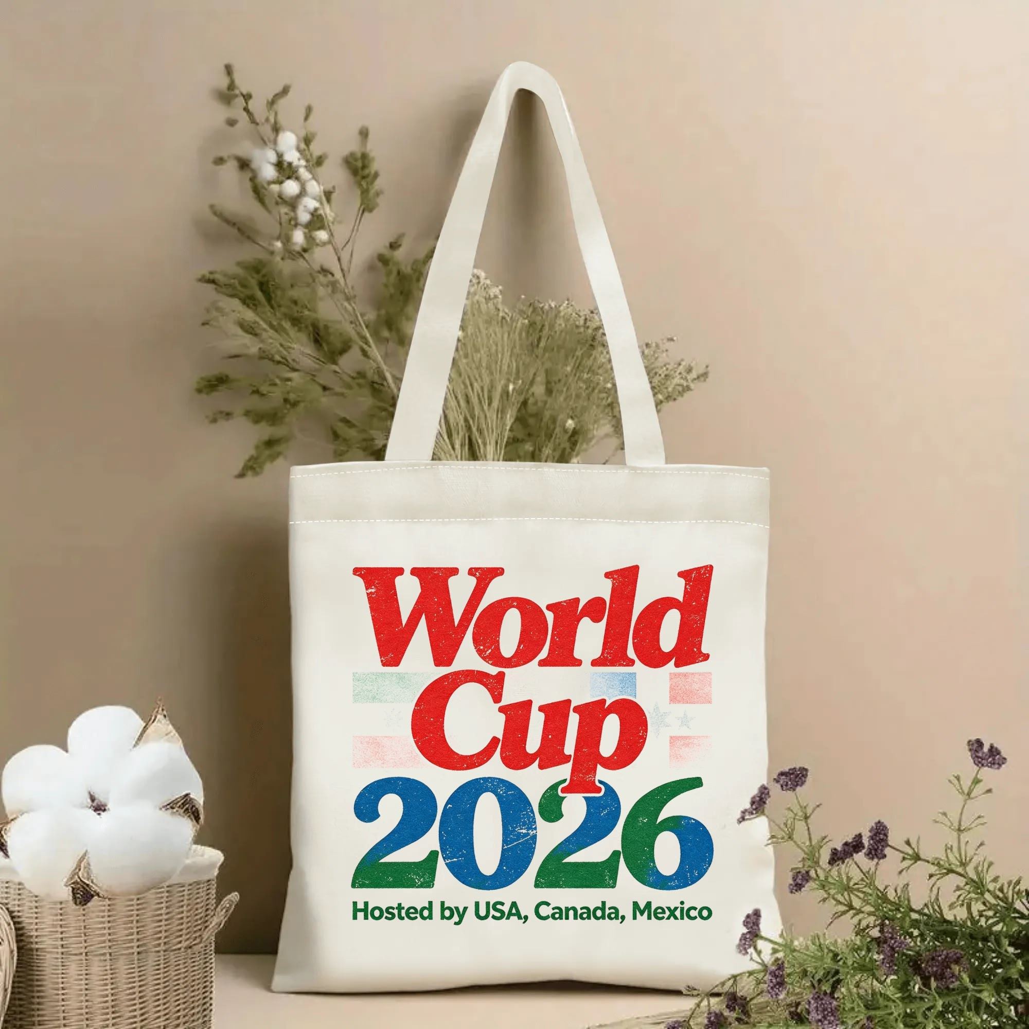 Lifestyle shot of the tote bag featuring a distressed "World Cup 2026" print in red, blue, and green, with the text "Hosted by USA, Canada, Mexico" below, displayed against a neutral background with plants.