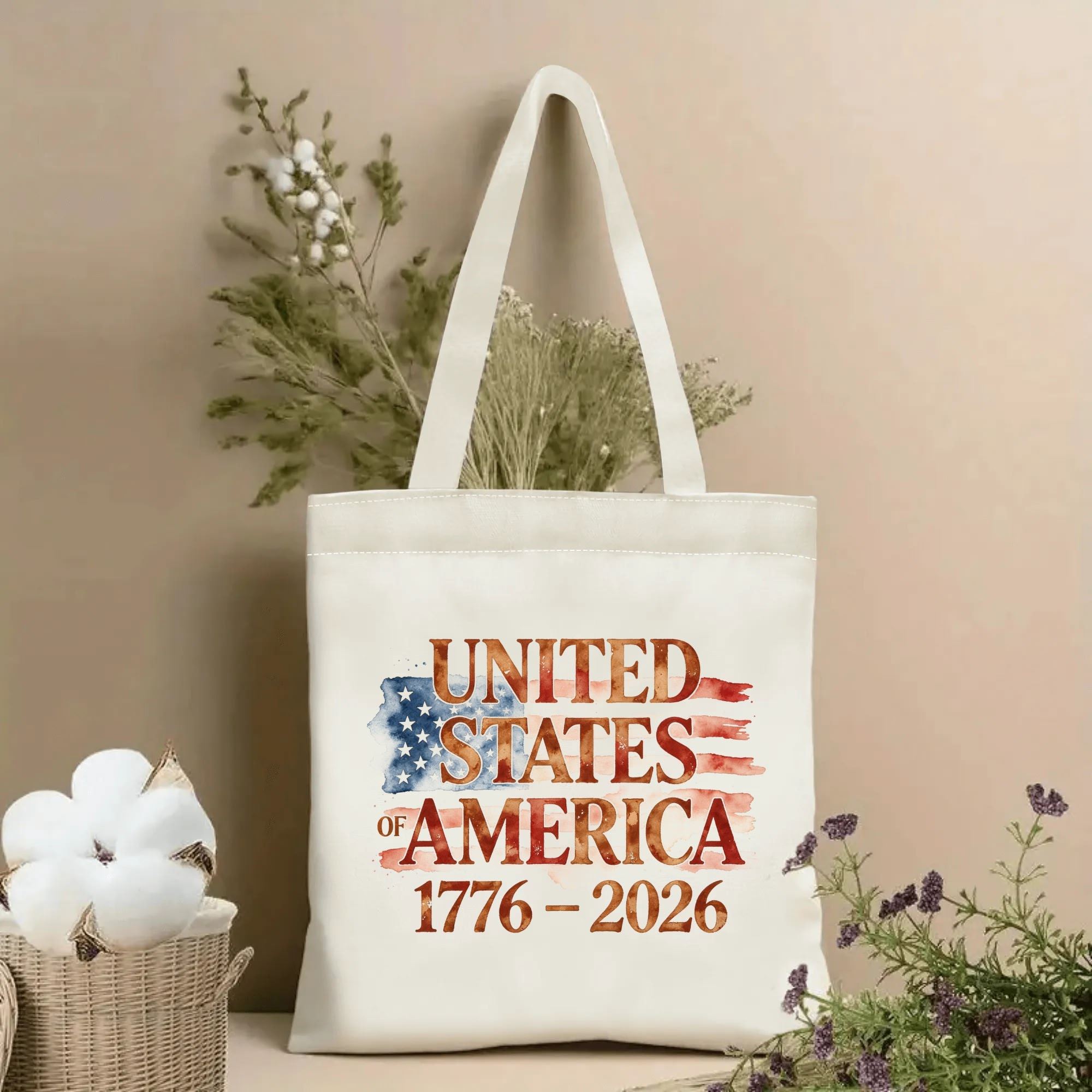 Lifestyle shot of the tote bag with a watercolor-style "UNITED STATES OF AMERICA 1776–2026" print, placed on a table with dried flowers.