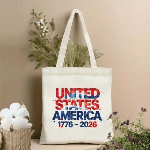 Lifestyle shot of the tote bag with a splatter-style "UNITED STATES OF AMERICA 1776–2026" print, placed on a table with dried flowers.