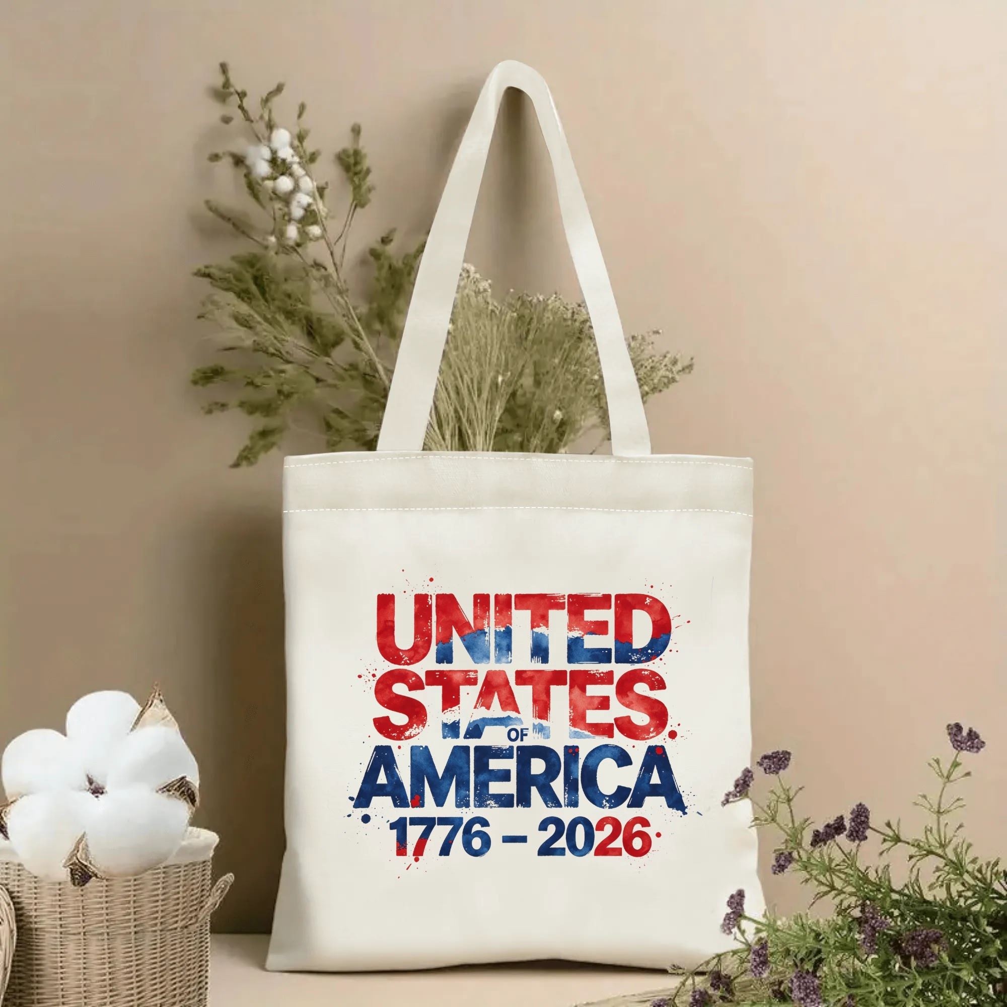Lifestyle shot of the tote bag with a splatter-style "UNITED STATES OF AMERICA 1776–2026" print, placed on a table with dried flowers.