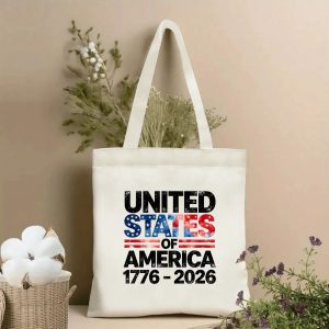 Lifestyle shot of the tote bag with a distressed "UNITED STATES OF AMERICA 1776–2026" print featuring flag-inspired stars and stripes, placed on a table with dried flowers.