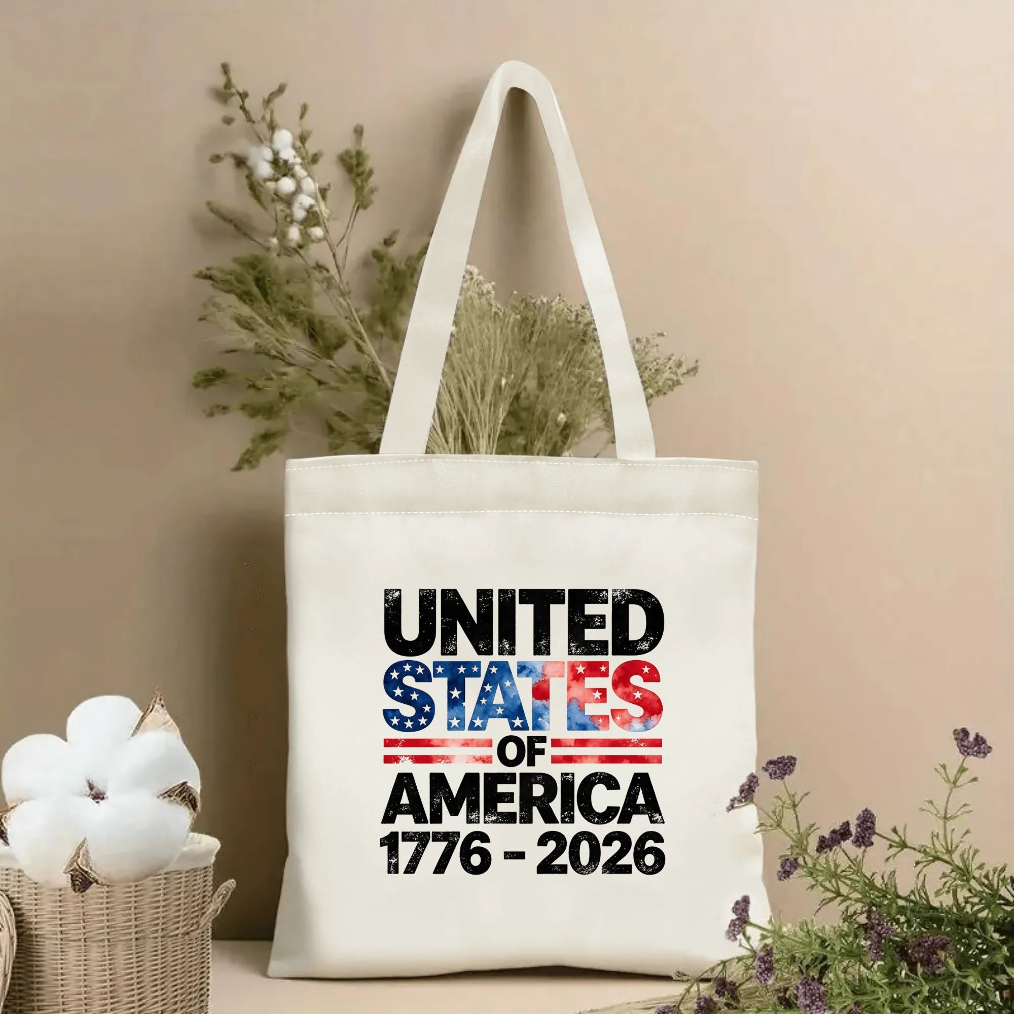 Lifestyle shot of the tote bag with a distressed "UNITED STATES OF AMERICA 1776–2026" print featuring flag-inspired stars and stripes, placed on a table with dried flowers.