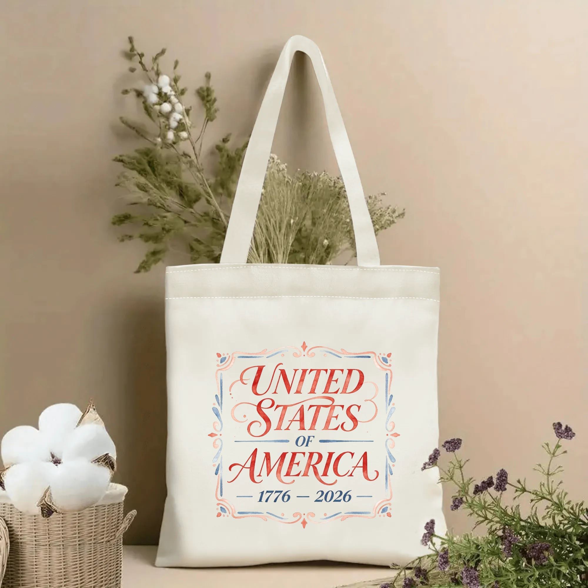 Lifestyle shot of the tote bag with an ornate watercolor-style "UNITED STATES OF AMERICA 1776–2026" print, placed on a table with dried flowers.