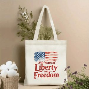 Lifestyle shot of the tote bag featuring a watercolor American flag print with "250 Years of Liberty and Freedom" text, styled with dried flowers.