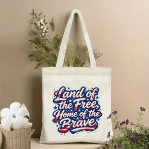 Lifestyle shot of the tote bag featuring a vibrant red-white-blue "Land of the Free, Home of the Brave" calligraphy print, styled with dried flowers.