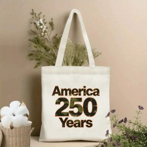 Lifestyle shot of the tote bag featuring a bold "America 250 Years" print in distressed camouflage-style lettering, styled with dried flowers.