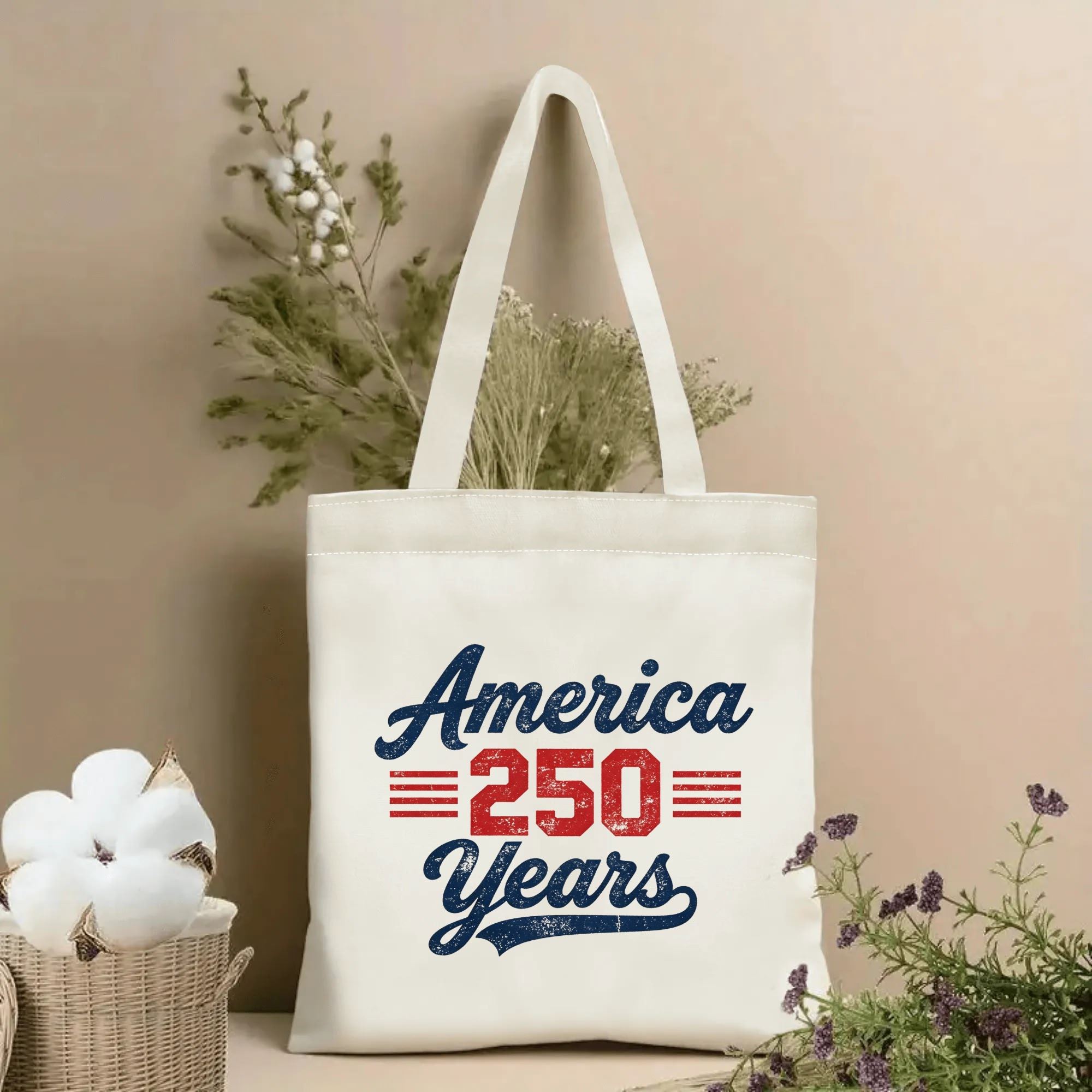 Lifestyle shot of the tote bag featuring a vintage script-style "America 250 Years" print in red-white-blue distressed lettering, styled with dried flowers.