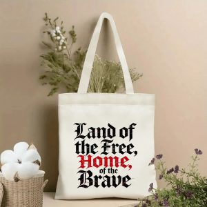 Lifestyle shot of the tote bag featuring a Gothic-style "Land of the Free, Home of the Brave" print with distressed red accents, styled with dried flowers.