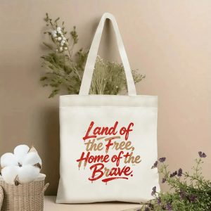 Lifestyle shot of the tote bag featuring a brushstroke-style "Land of the Free, Home of the Brave" print in red and gold with paint drip details, styled with dried flowers.