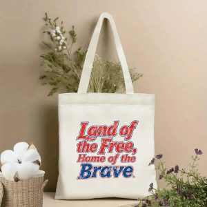 Lifestyle shot of the tote bag featuring a retro varsity-style "Land of the Free, Home of the Brave" print in red, white, and blue with distressed details, styled with dried flowers.