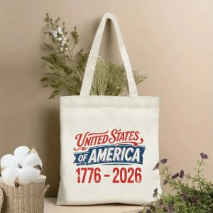 Lifestyle shot of the tote bag featuring a vintage "United States of America 1776-2026" print in red, white, and blue distressed lettering, styled with dried flowers.