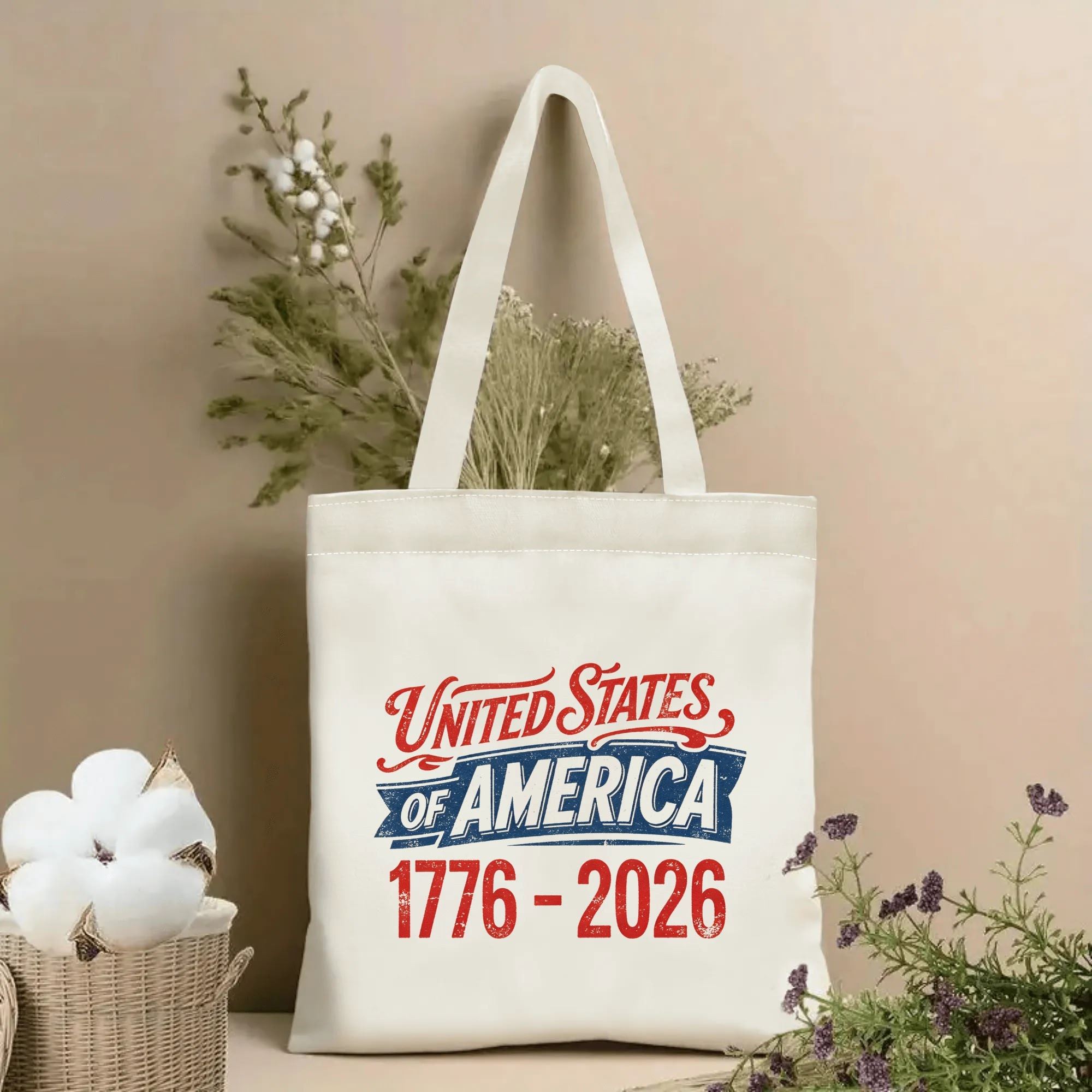 Lifestyle shot of the tote bag featuring a vintage "United States of America 1776-2026" print in red, white, and blue distressed lettering, styled with dried flowers.