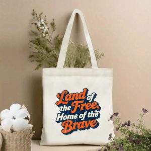 Lifestyle shot of the tote bag featuring a retro 70s-style "Land of the Free, Home of the Brave" print in orange, navy, and cream with cracked distressed details, styled with dried flowers.