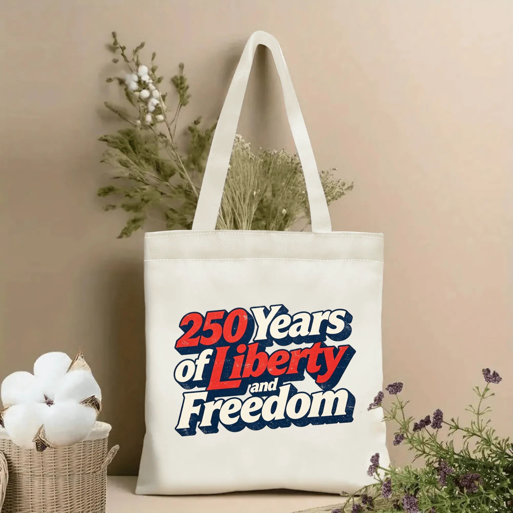 Lifestyle shot of the tote bag featuring a retro 3D-style "250 Years of Liberty and Freedom" print in red, white, and blue distressed lettering, styled with dried flowers.