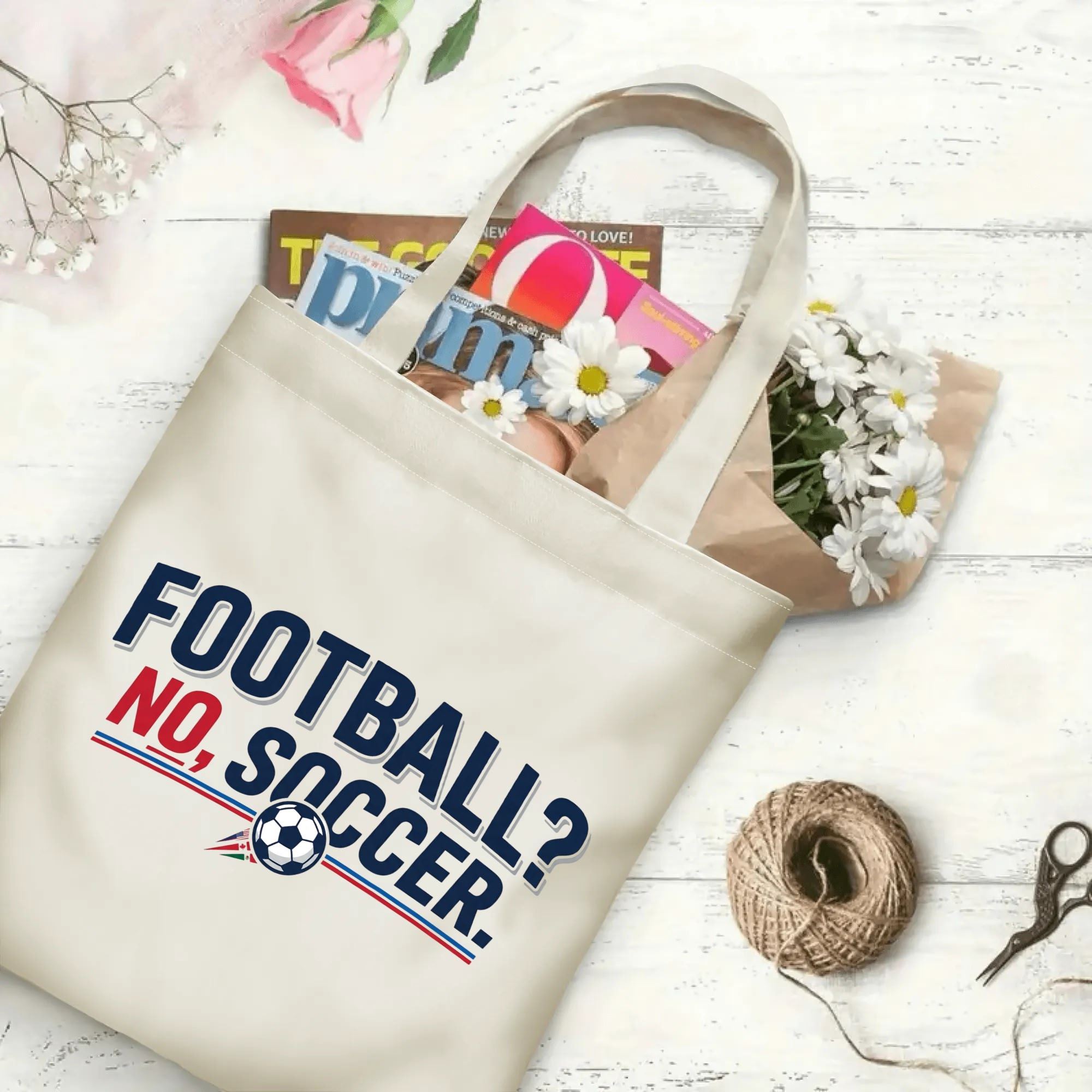 Clean two-tone soccer slogan graphic canvas tote bag laid flat with magazines and daisies, reusable minimalist sport fan daily shopping bag