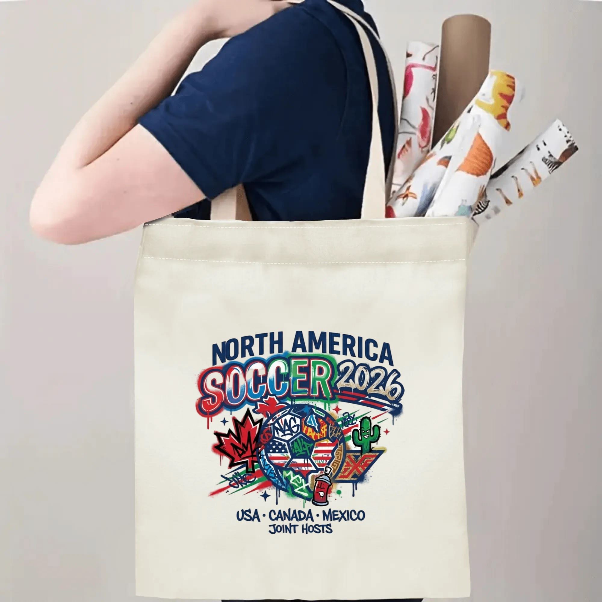 Person slinging 2026 USA Canada Mexico graffiti soccer badge canvas tote bag over shoulder, large capacity urban-style football supporter carry bag for stadium trips