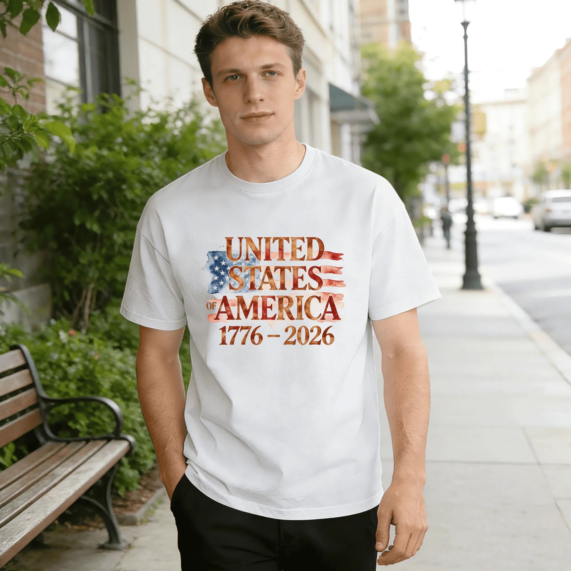 Lifestyle shot of a man wearing the white “USA 1776-2026” watercolor flag T-shirt, outdoors in an urban setting