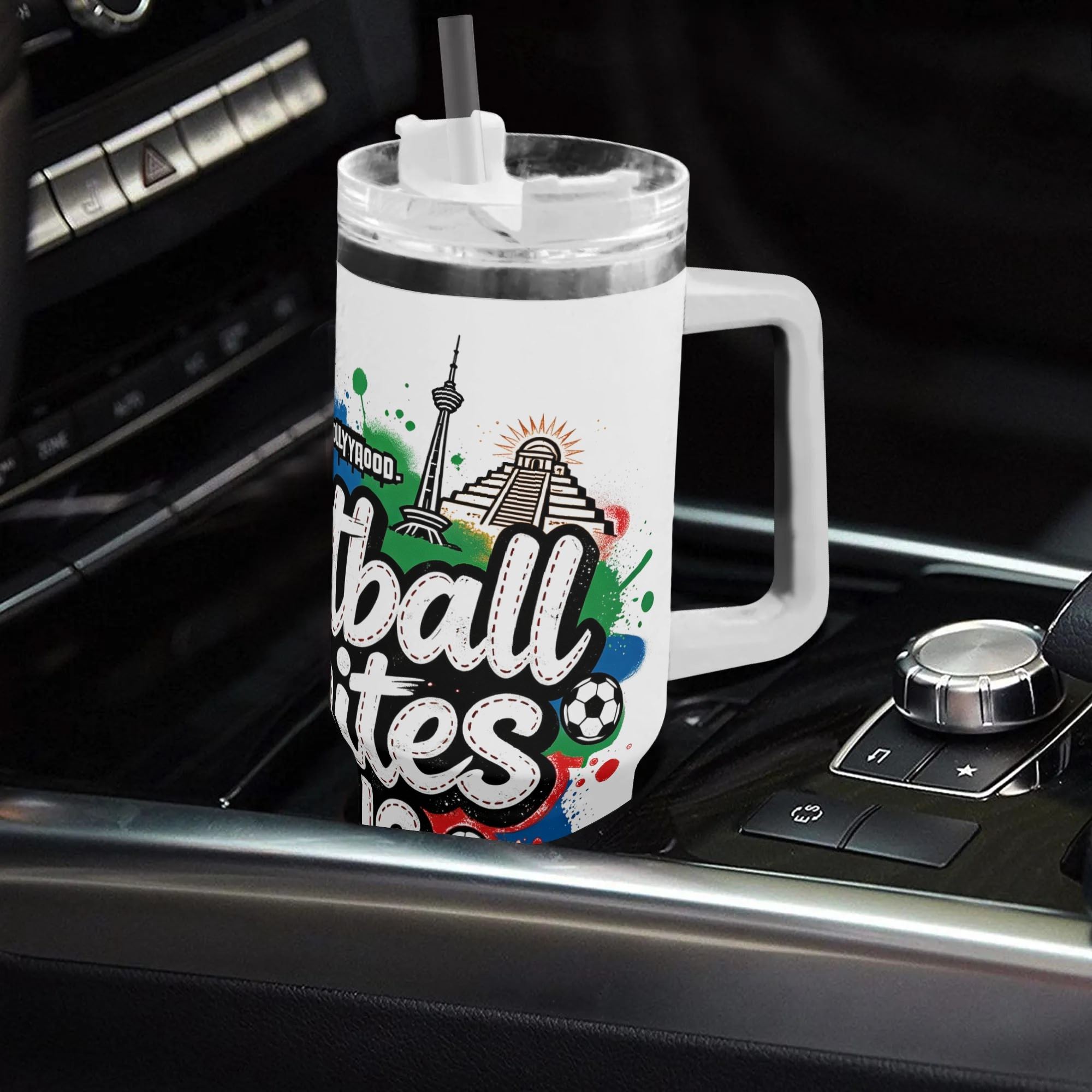 White 40oz insulated tumbler in a car cup holder, featuring a graffiti-style "Football Unites Us" print with North American landmarks (Statue of Liberty, Hollywood sign, CN Tower, Chichen Itza) and soccer ball details.