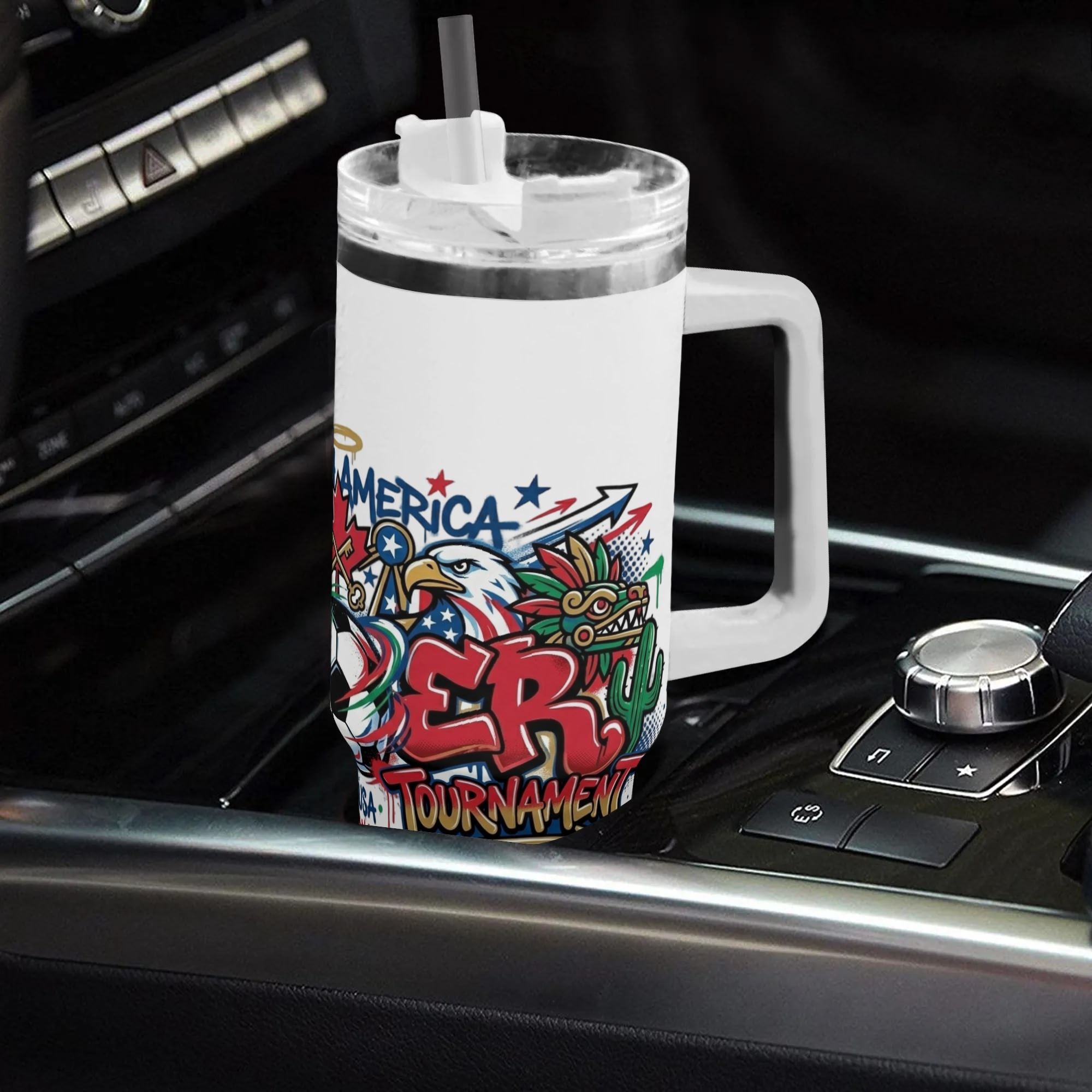 White 40oz handled insulated tumbler with eagle quetzal graffiti soccer graphic, placed on car gear shift console, 2026 World Cup fan stainless steel travel mug for driving